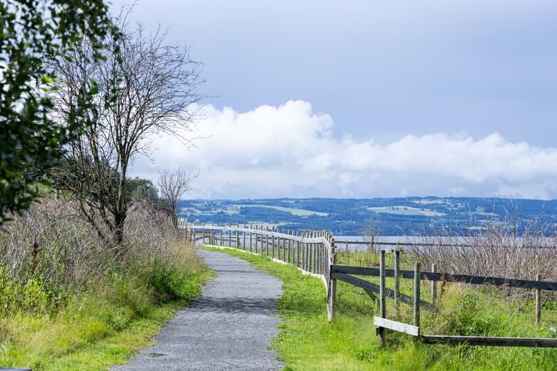 Grusvei langs et gjerde, med utsyn over Mjøsa og åsene bortenfor.